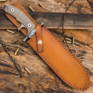 May include: A tan and silver hunting knife with a textured handle is partially sheathed in a brown leather case. The case is stitched and has a strap with a brass button. Several bullets and a rope are scattered around the knife.