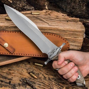 May include: A large knife with a silver blade and a tan and brown handle. The knife is held in a hand, with a brown leather sheath in the background. The knife has a wide blade and a guard. Several bullets are scattered around the knife.