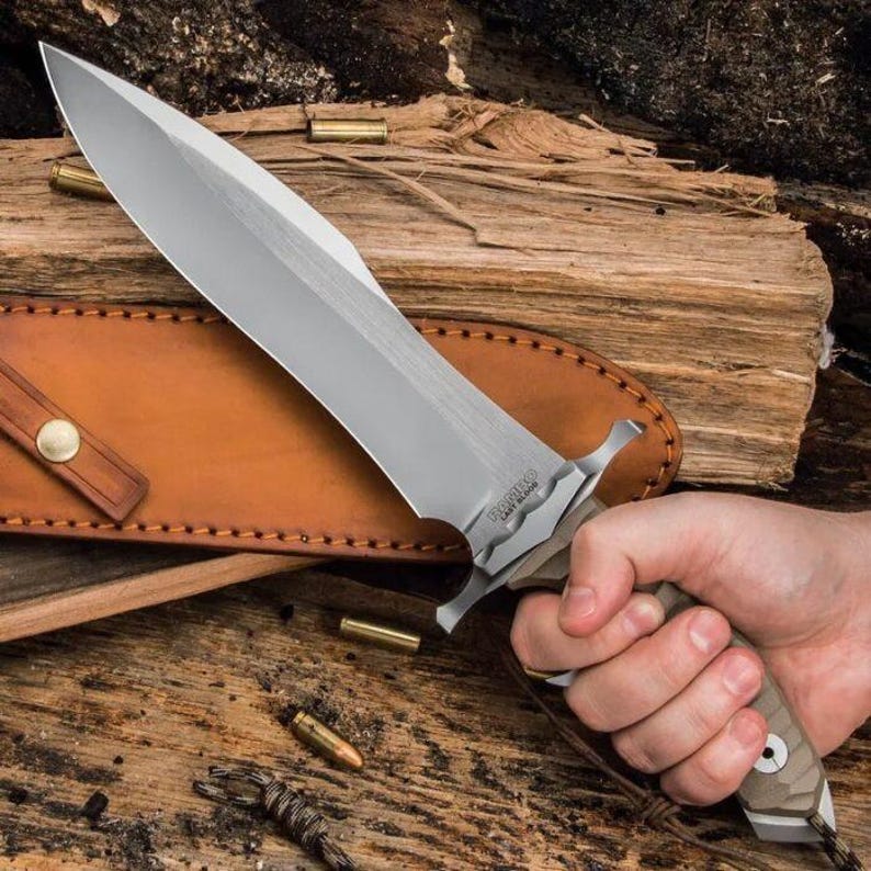 May include: A large knife with a silver blade and a tan and brown handle. The knife is held in a hand, with a brown leather sheath in the background. The knife has a wide blade and a guard. Several bullets are scattered around the knife.