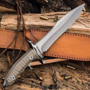 May include: A large, silver-bladed knife with a tan handle and a leather sheath. The knife has a double-edged blade and a textured handle. Several bullets are scattered around the knife on a wooden surface.