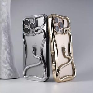 May include: Two smartphone cases, one in silver and one in gold, each featuring a three-dimensional metallic snake design that wraps around the camera module and extends down the back of the case, showcasing a textured, scale-like surface finish.