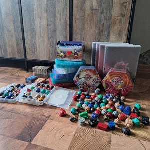 Op de afbeelding: Een verzameling Bakugan-speelgoed en accessoires, waaronder karakterballen, opbergdozen en speelkaarten. Het speelgoed is in verschillende kleuren en ontwerpen, verspreid over een houten vloer.