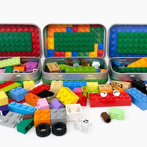 May include: Three open metal tins with colorful building block bases, each filled with assorted blocks. Blocks are red, yellow, blue, green, orange, and gray. Some blocks are scattered in front of the tins, including a red block with white eyes.