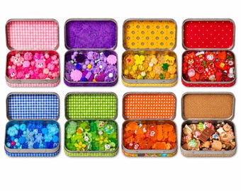 May include: Eight rectangular metal tins, each filled with small, colorful plastic charms and beads. The interior lids feature distinct patterns including gingham, polka dots, and floral prints in pink, purple, yellow, red, blue, green, orange, and tan.