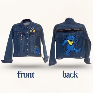 May include: A denim jacket with front and back views. The front features a blue flower patch, pins, and a small yellow patch. The back displays a blue bear patch with a yellow collar and black trim. The jacket is a medium wash.