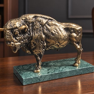 May include: A bronze-toned metal sculpture of a bison standing in a profile position. The animal figure features a textured, weathered surface and is mounted on a rectangular, polished green marble base with visible dark veining throughout.