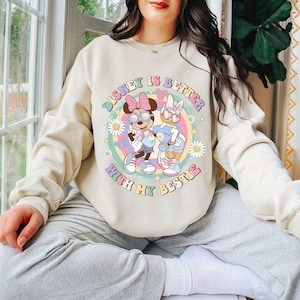 May include: Beige sweatshirt featuring a colorful graphic of Minnie Mouse and Daisy Duck with the text "Disney is Better With My Bestie". The design includes a rainbow, flowers, and stars.