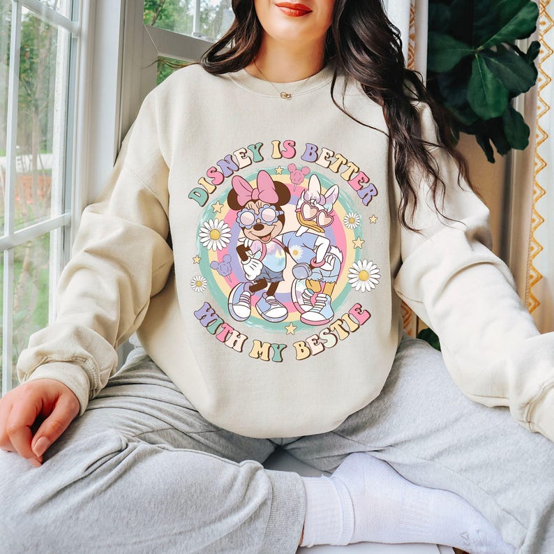May include: Beige sweatshirt featuring a colorful graphic of Minnie Mouse and Daisy Duck with the text "Disney is Better With My Bestie". The design includes a rainbow, flowers, and stars.