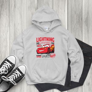 May include: A light gray hoodie featuring a graphic of Lightning McQueen from the movie Cars. The graphic includes the text "LIGHTNING MCQUEEN" and "ULTIMATE SPEED Think Fast Rusteze" with a red race car design. Black sneakers and dark jeans are also visible.
