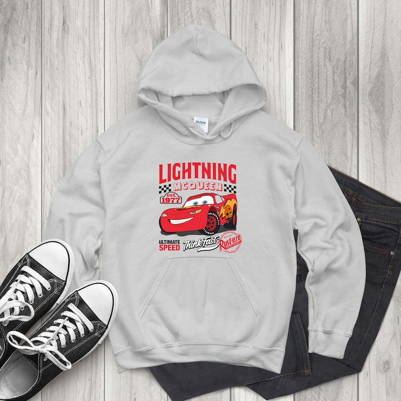 May include: A light gray hoodie featuring a graphic of Lightning McQueen from the movie Cars. The graphic includes the text "LIGHTNING MCQUEEN" and "ULTIMATE SPEED Think Fast Rusteze" with a red race car design. Black sneakers and dark jeans are also visible.