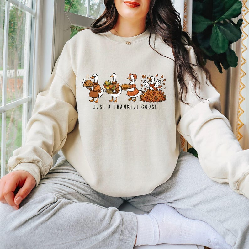 Puede incluir: Sudadera de color crema con un gr&aacute;fico de cuatro gansos de dibujos animados vestidos para Acci&oacute;n de Gracias. Los gansos llevan comida y est&aacute;n de pie en un mont&oacute;n de hojas. El texto "JUST A THANKFUL GOOSE" est&aacute; impreso debajo.