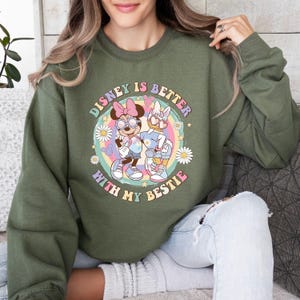 May include: Olive green sweatshirt with the text "Disney is Better With My Bestie" in a circular design. The design features Minnie Mouse and Daisy Duck in a colorful, retro style. The sweatshirt is a casual, comfortable garment.