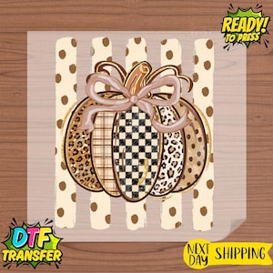 May include: A DTF transfer sheet featuring a pumpkin graphic with a light brown bow. The pumpkin has segments with leopard print, plaid, and black and white checkerboard patterns. The background consists of vertical cream stripes with brown dots.