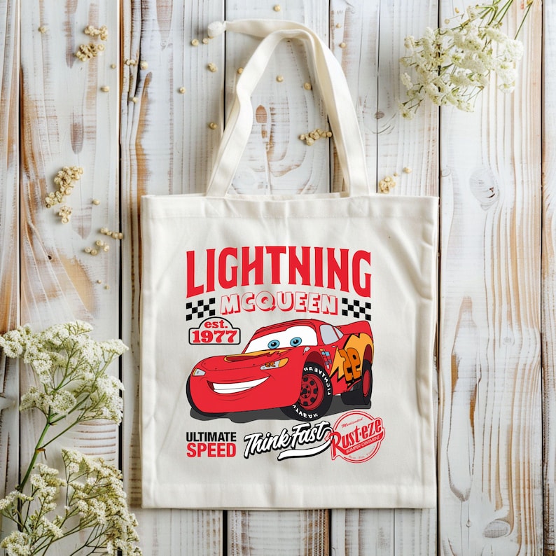 May include: Off-white canvas tote bag featuring a red Lightning McQueen race car graphic. The design includes the text "LIGHTNING MCQUEEN" in red, with "est. 1977" and "ULTIMATE SPEED Think Fast Rust-eze" also printed on the bag.