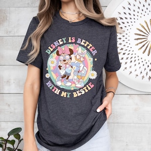 May include: Dark gray t-shirt featuring a colorful graphic of Minnie Mouse and Daisy Duck wearing sunglasses. The text "DISNEY IS BETTER WITH MY BESTIE" is printed in a rainbow circle around the characters. Daisies and stars surround the characters.