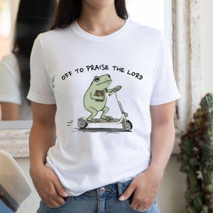 May include: White t-shirt featuring a cartoon frog riding a scooter and holding a book. The text "OFF TO PRAISE THE LORD" is printed above the frog. The frog is light green, and the scooter is white and gray.