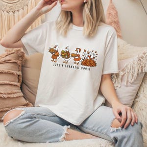 Puede incluir: Camiseta de color crema con una ilustraci&oacute;n de Acci&oacute;n de Gracias. La ilustraci&oacute;n muestra gansos con un pavo, cestas de comida y hojas. El texto "JUST A THANKFUL GOOSE" est&aacute; impreso debajo. El dise&ntilde;o es en colores oto&ntilde;ales.