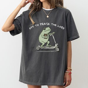 May include: Dark gray t-shirt featuring a cartoon frog riding a scooter and holding a book. The text "OFF TO PRAISE THE LORD" is arched above the frog. The shirt has a relaxed fit and short sleeves.