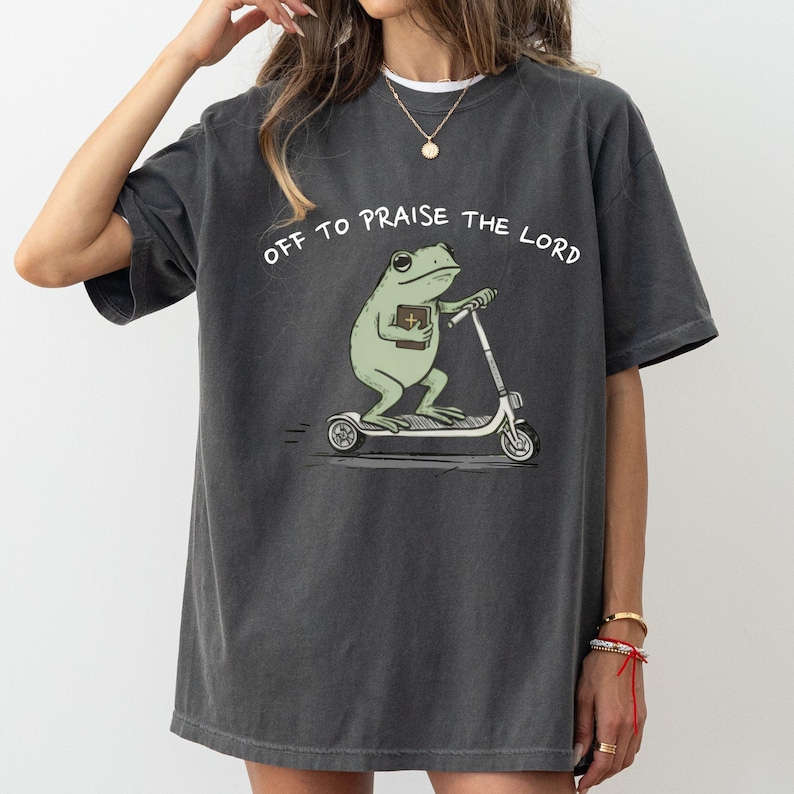 May include: Dark gray t-shirt featuring a cartoon frog riding a scooter and holding a book. The text "OFF TO PRAISE THE LORD" is arched above the frog. The shirt has a relaxed fit and short sleeves.