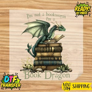 May include: A DTF transfer sheet featuring a green dragon perched atop a stack of vintage books. The text reads, "I'm not a bookworm I'm a Book Dragon." The design includes small green plants and rocks at the base of the book stack.