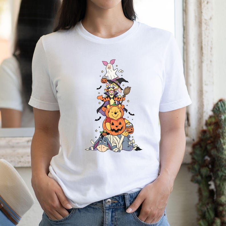 Op de afbeelding: Een wit t-shirt met korte mouwen en een verticale afbeelding van gestapelde stripfiguren in Halloween-kostuums, waaronder een geest, een heks, een tijger, een beer in een pompoen en een ezel in verband met vleermuizen.