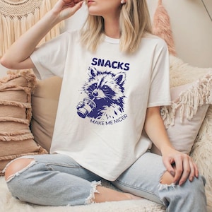 May include: Cream-coloured t-shirt featuring a navy blue raccoon graphic drinking from a cup. The text "SNACKS MAKE ME NICER" is above and below the raccoon illustration. A casual, everyday wear item.