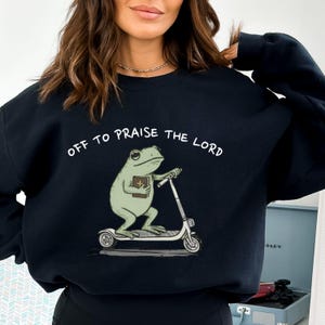 May include: Black sweatshirt with the text "OFF TO PRAISE THE LORD" in white. The graphic features a green frog riding a scooter while holding a book. The frog has large eyes and is wearing a cross. The scooter is white with black wheels.