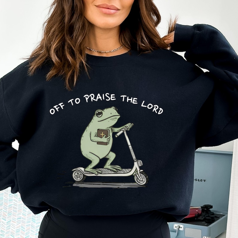 May include: Black sweatshirt with the text "OFF TO PRAISE THE LORD" in white. The graphic features a green frog riding a scooter while holding a book. The frog has large eyes and is wearing a cross. The scooter is white with black wheels.