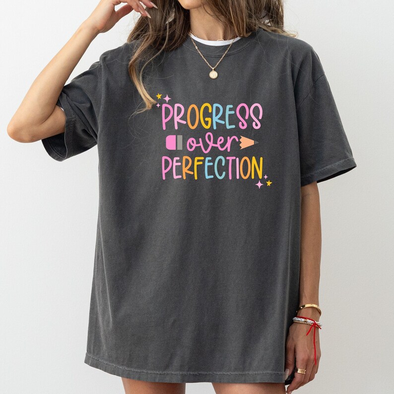 May include: Dark gray t-shirt with the text "PROGRESS over PERFECTION" in colorful, playful lettering. The design includes a pink eraser, a yellow pencil, and small star accents. The shirt is a casual, oversized style.