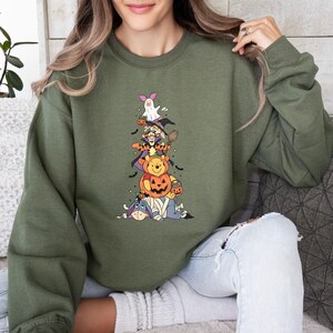 Op de afbeelding: Een olijfgroen sweatshirt met ronde hals en een grafische print van gestapelde stripfiguren verkleed voor Halloween. De stapel bevat Iejoor als mummie, Winnie de Poeh in een pompoen, Teigetje als tovenaar en Knorretje als spookje.