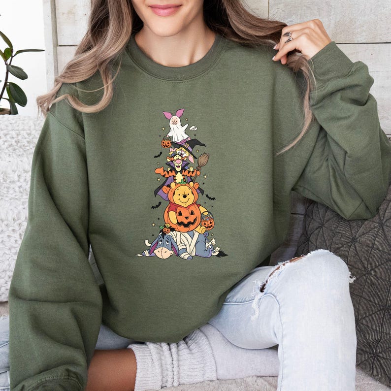 Op de afbeelding: Een olijfgroen sweatshirt met ronde hals en een grafische print van gestapelde stripfiguren verkleed voor Halloween. De stapel bevat Iejoor als mummie, Winnie de Poeh in een pompoen, Teigetje als tovenaar en Knorretje als spookje.