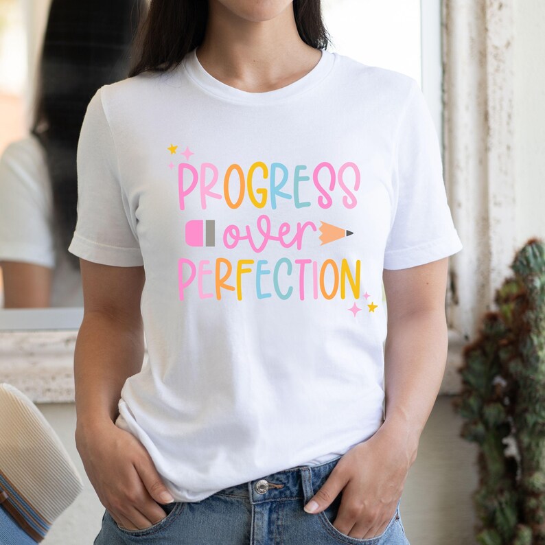May include: White t-shirt with the phrase "PROGRESS over PERFECTION" in colorful bubble letters. A pink pencil and yellow stars accent the design. The shirt is a casual, everyday wear item.