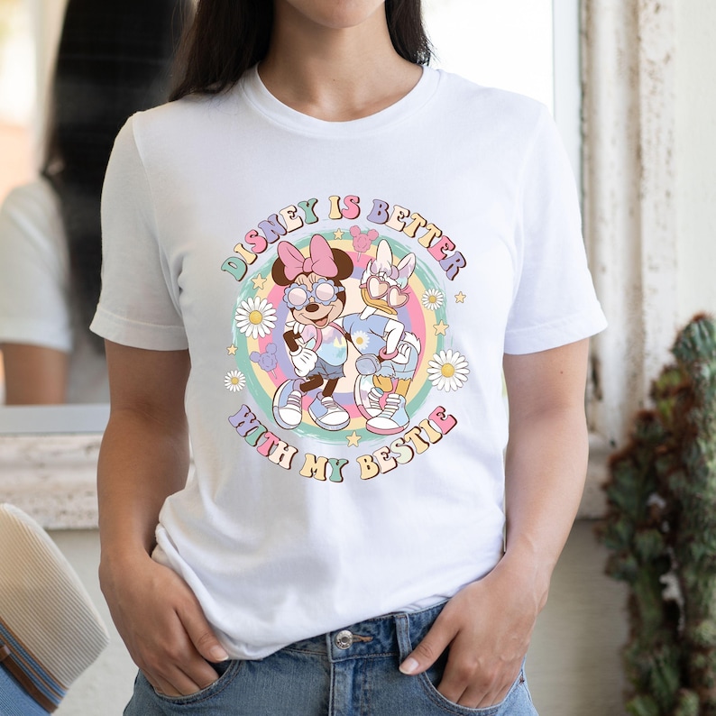 May include: White t-shirt featuring a colorful graphic of Minnie Mouse and Daisy Duck with the text "Disney is Better With My Bestie." The design includes pastel colors, stars, and daisies, creating a cheerful and playful aesthetic.