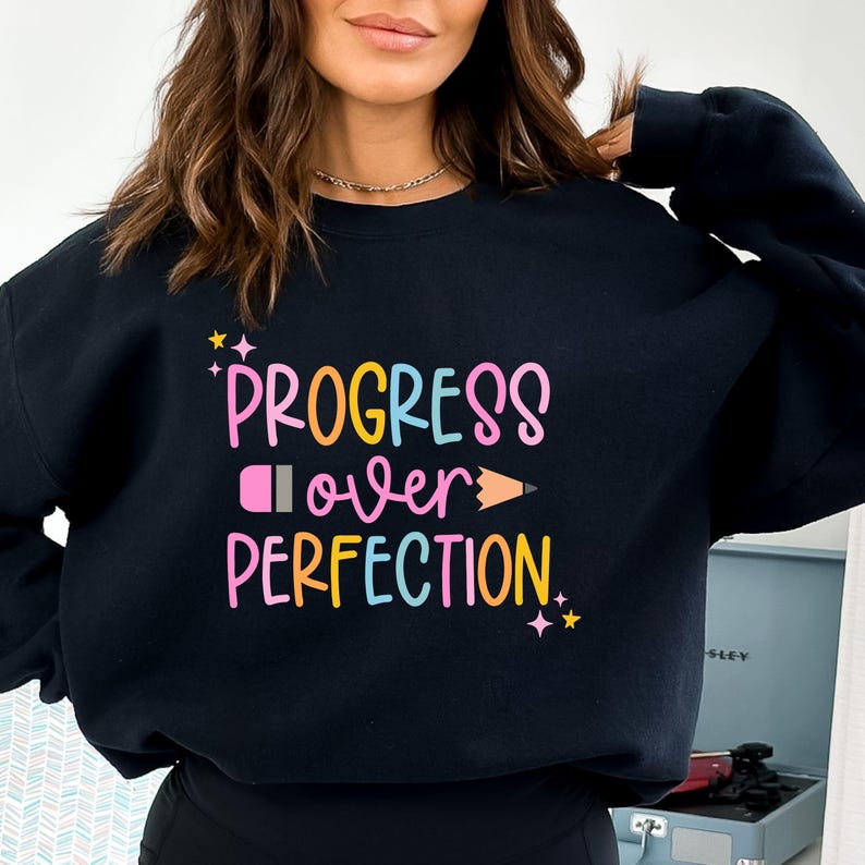 May include: Black sweatshirt with the text "PROGRESS over PERFECTION" in colorful bubble letters. The design includes a pink eraser and an orange pencil. The sweatshirt has a crew neck.