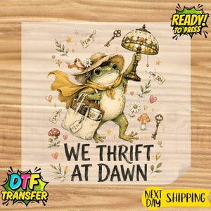May include: A graphic print featuring a green frog wearing a straw hat and yellow cape, holding a stained glass lamp and a tote bag. The design includes small price tags, keys, mushrooms, and the text WE THRIFT AT DAWN in black lettering.