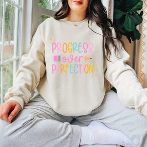 May include: Beige sweatshirt with the text "PROGRESS over PERFECTION" in colorful letters. The design includes a pencil and stars. The sweatshirt is long-sleeved and has a crew neck.