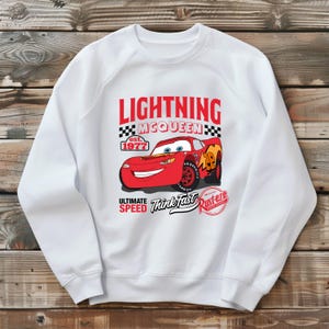 May include: White sweatshirt featuring a graphic of Lightning McQueen, a red race car from the movie Cars. The design includes the text "LIGHTNING McQUEEN" above the car, with "est. 1977" and "ULTIMATE SPEED Think Fast Rust-eze" below.
