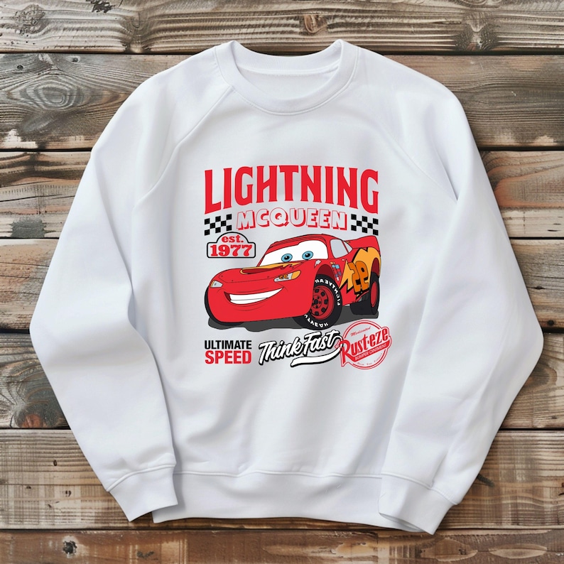 May include: White sweatshirt featuring a graphic of Lightning McQueen, a red race car from the movie Cars. The design includes the text "LIGHTNING McQUEEN" above the car, with "est. 1977" and "ULTIMATE SPEED Think Fast Rust-eze" below.