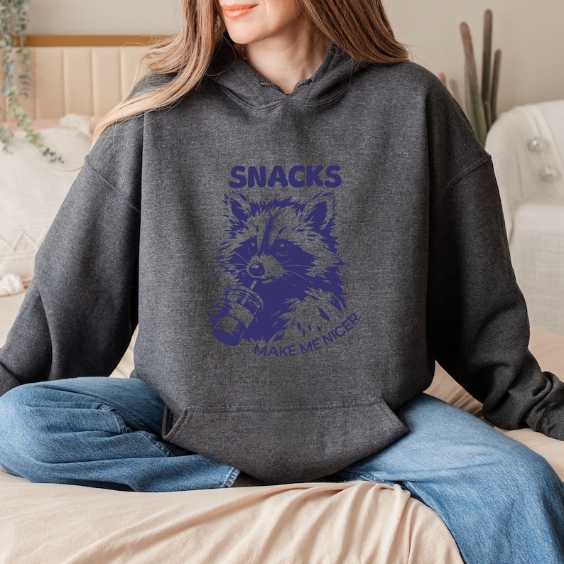 May include: A charcoal grey hoodie with a purple raccoon illustration and the text "SNACKS" and "MAKE ME NICER". The raccoon is depicted with a drink. The hoodie has a front pocket and a drawstring hood, typical of casual wear.