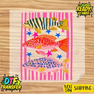 May include: DTF transfer featuring three colorful fish illustrations. The top fish has green and yellow stripes, the middle fish is red and orange, and the bottom fish is pink with blue spots. Pink vertical stripes and blue and pink stars are also present.