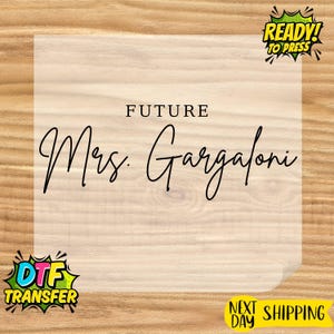 May include: A light-colored transfer sheet with the words "FUTURE Mrs. Gargaloni" in a cursive font. The words are centered on the sheet. The background is a light wood grain. The words "READY! TO PRESS" and "DTF TRANSFER" are in the corners.