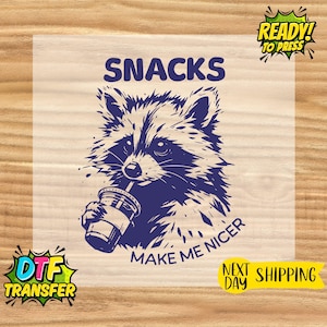 May include: A navy blue graphic of a raccoon drinking from a cup, with the words "SNACKS" and "MAKE ME NICER". The design is ready to press and includes "DTF TRANSFER" and "NEXT DAY SHIPPING" text.