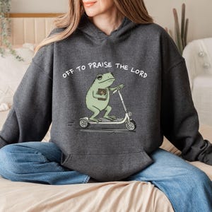 May include: Dark gray hoodie with the text "OFF TO PRAISE THE LORD." The hoodie features a cartoon frog riding a scooter and holding a book. The frog is light green, and the scooter is white.