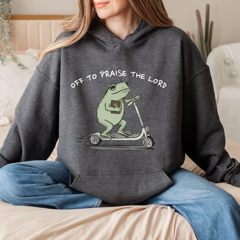 May include: Dark gray hoodie with the text "OFF TO PRAISE THE LORD." The hoodie features a cartoon frog riding a scooter and holding a book. The frog is light green, and the scooter is white.