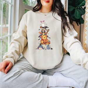 Op de afbeelding: Een cr&egrave;mekleurig sweatshirt met ronde hals en een centrale afbeelding van gestapelde stripfiguren in Halloween-kostuums, waaronder een geest, een heks, een pompoen en een mummie, geaccentueerd met kleine zwarte vleermuizen en sterren.