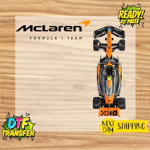May include: A top-down view of a McLaren Formula 1 race car. The car is primarily orange and black with the number 4 on the side. The image includes the McLaren logo and the text "READY! TO PRESS".