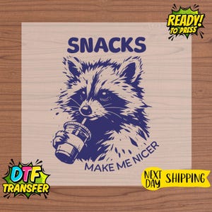 May include: A navy blue graphic of a raccoon drinking from a cup, with the words "SNACKS" and "MAKE ME NICER". The image is a DTF transfer, ready to press, with next-day shipping.