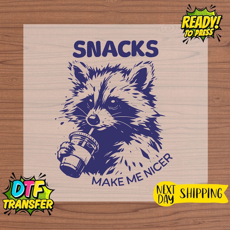 May include: A navy blue graphic of a raccoon drinking from a cup, with the words "SNACKS" and "MAKE ME NICER". The image is a DTF transfer, ready to press, with next-day shipping.