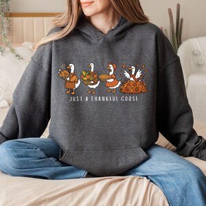 Puede incluir: Sudadera con capucha gris oscuro con un gr&aacute;fico de cuatro gansos de dibujos animados, cada uno con elementos tem&aacute;ticos de Acci&oacute;n de Gracias. El texto "JUST A THANKFUL GOOSE" est&aacute; debajo. La sudadera se combina con unos vaqueros azules.