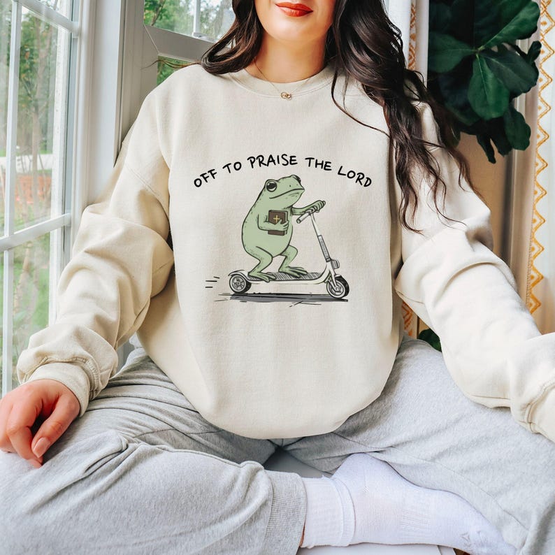 May include: Cream-colored sweatshirt with the text "OFF TO PRAISE THE LORD" above a cartoon frog riding a scooter and holding a book. The frog is light green, and the scooter is gray.